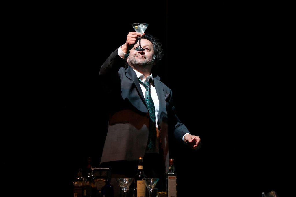 Actor en traje oscuro ajustándose las gafas, en postura teatral sobre escenario oscuro