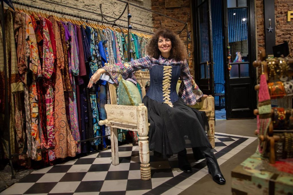 Mujer con cabello rizado sentada en silla blanca entre prendas de colores en tienda de ropa vintage