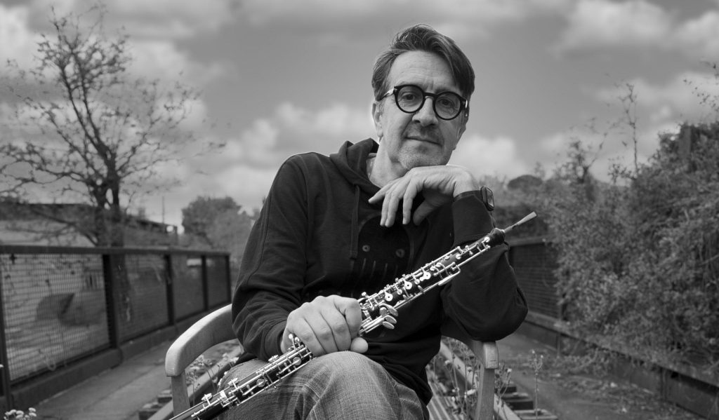 Gianfranco Bortolato, oboísta italiano, sostiene su instrumento en primer plano frente a paisaje urbano gris