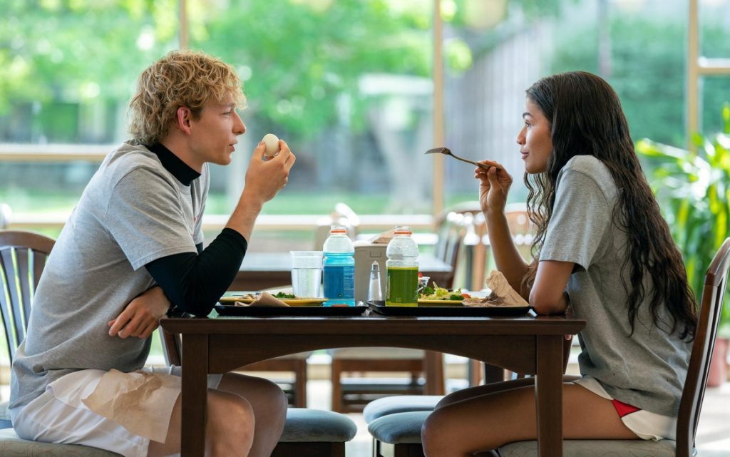 Dos personajes comparten una conversación íntima en una mesa durante el almuerzo, con plantas verdes de fondo