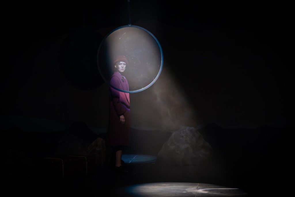 Performer en escena oscura sosteniendo gran esfera luminosa con luz dramática y polvo en suspenso