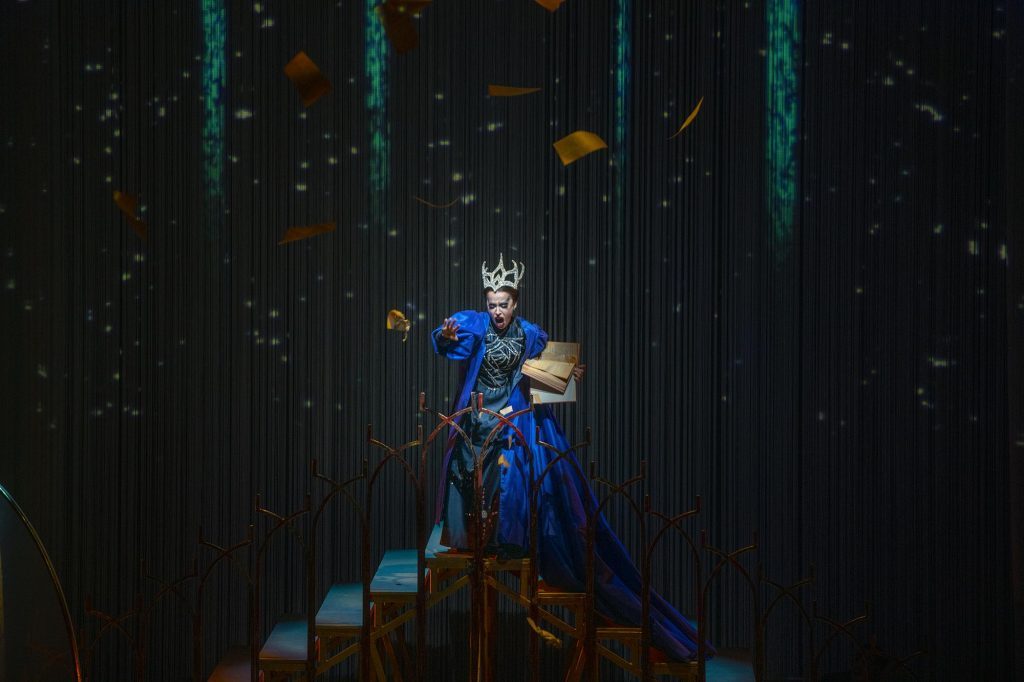 Reina de la Noche en traje azul y dorado sobre escaleras, iluminada dramáticamente contra fondo oscuro con efecto