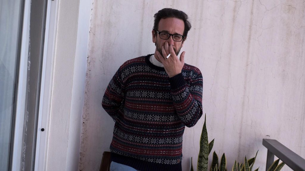 Hombre con gafas y suéter tejido de colores oscuros junto a una planta, en interior con pared blanca
