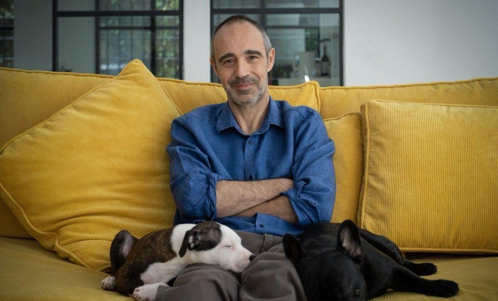Niccolò Ammaniti sentado en sofá amarillo con gatos negros, brazos cruzados, fondo de ventanas