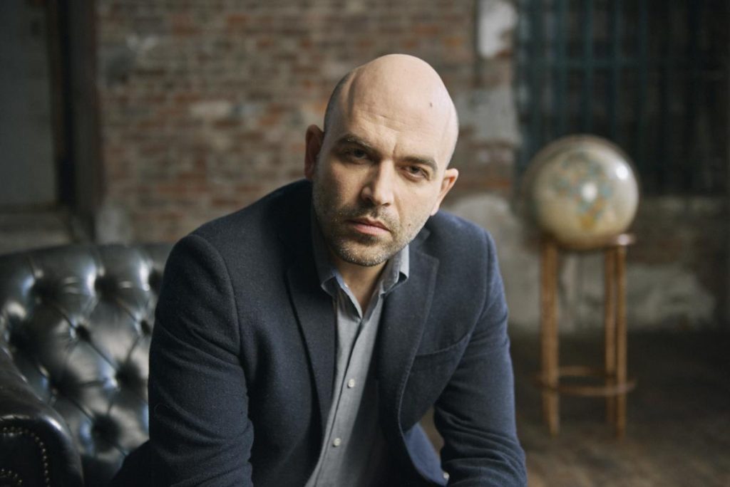Roberto Saviano, escritor italiano, en estudio con traje azul marino y fondo de ladrillo