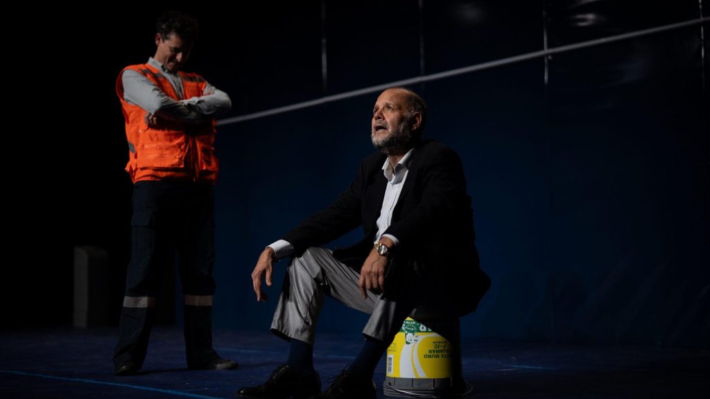 Dos actores en escena teatral: uno de pie con chaleco naranja sosteniendo una cuerda, otro sentado en jeans frente