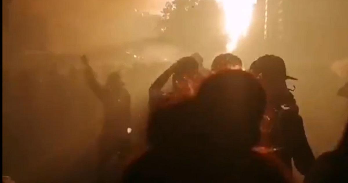 Multitud de personas envueltas en humo denso durante manifestación nocturna, con luces brillantes de fondo iluminando