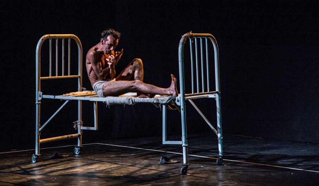 Actor sobre cama metálica en escena teatral, representando la angustia y transformación del protagonista