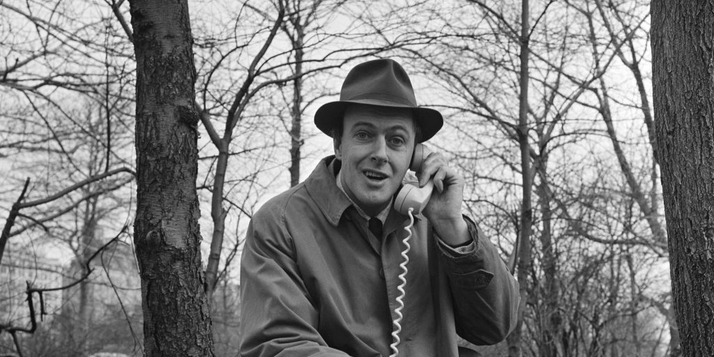 Roald Dahl con sombrero y abrigo, apoyado en un árbol desnudo, en retrato en blanco y negro