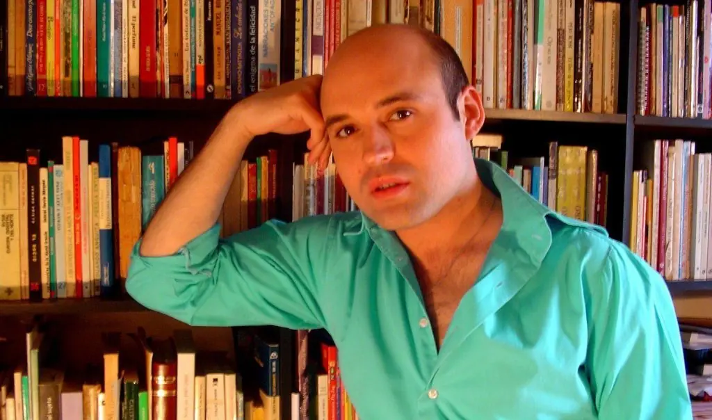 Hombre de camisa verde claro apoyando cabeza en mano, sonriendo ante estantería de libros de colores variados