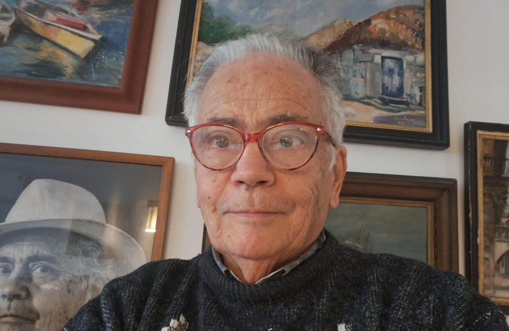 Edmundo Moure Rojas, escritor chileno, retratado en primer plano con gafas y expresión serena frente a cuadros de fondo