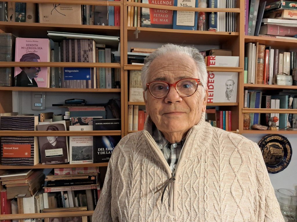 Edmundo Moure Rojas frente a estantería con libros, vistiendo suéter gris y lentes