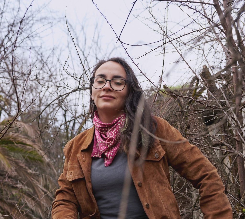Mujer con lentes y bandana roja, abrigo marrón, entre ramas desnudas en ambiente otoñal