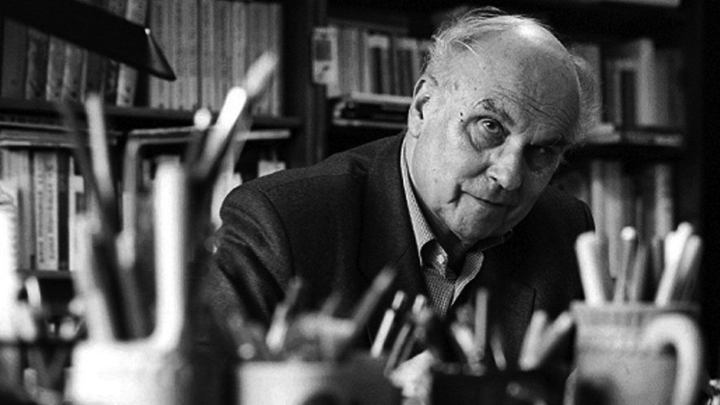 Ryszard Kapuscinski en su escritorio, rodeado de objetos y libros, mirando hacia la cámara en fotografía en blanco