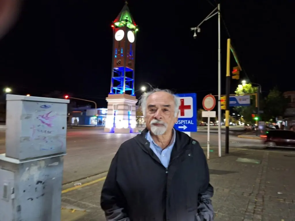 Juan Mihovilovich frente a reloj iluminado de noche, plaza urbana con arquitectura histórica de fondo