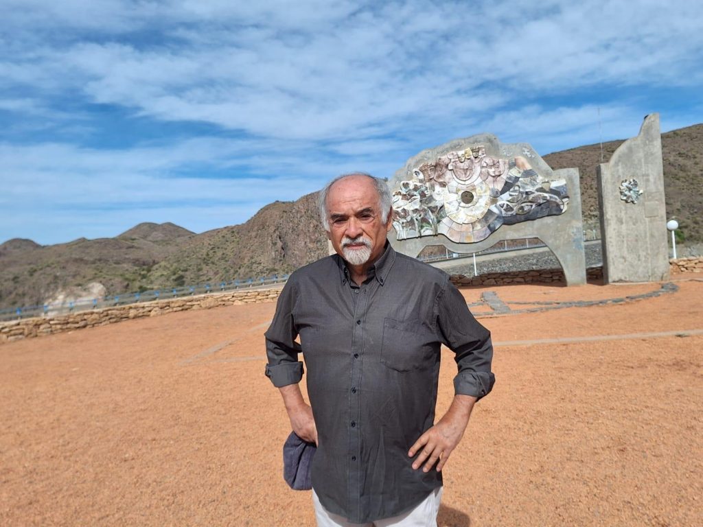 Hombre de mediana edad en camisa gris oscura posando en terreno árido con montaña nevada al fondo, Juan Mihovilovich