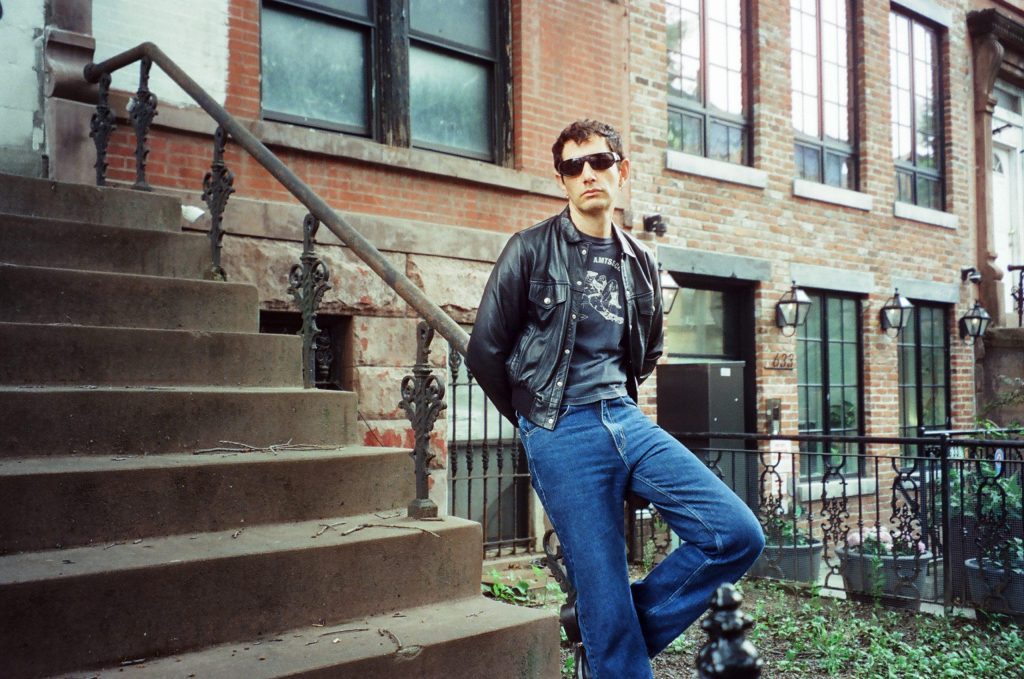 Álex Anwandter posando en escaleras de ladrillo rojo, con jeans y chaqueta oscura, gafas de sol