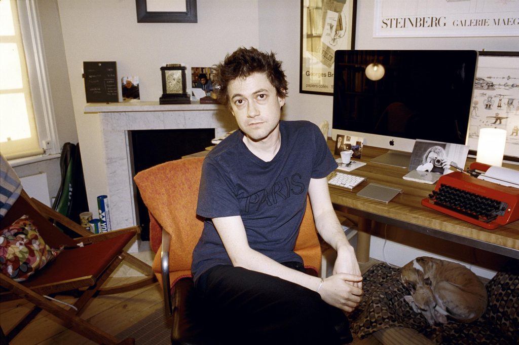 Adam Thirlwell sentado en escritorio con computadora, rodeado de libros y objetos personales en su estudio