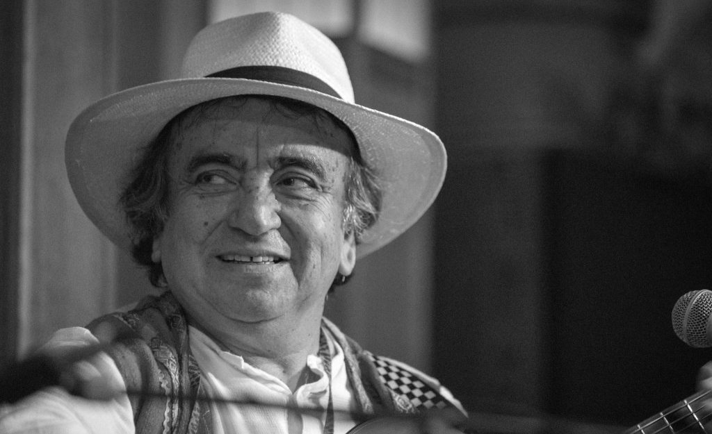 Hombre con sombrero de ala ancha y camisa de cuadros sonríe hacia la cámara en fotografía en blanco y negro