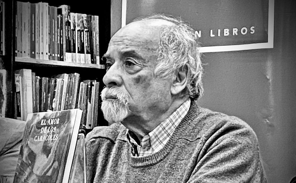 Juan Mihovilovich, escritor chileno de cabello blanco y bigote, en biblioteca con libros de fondo