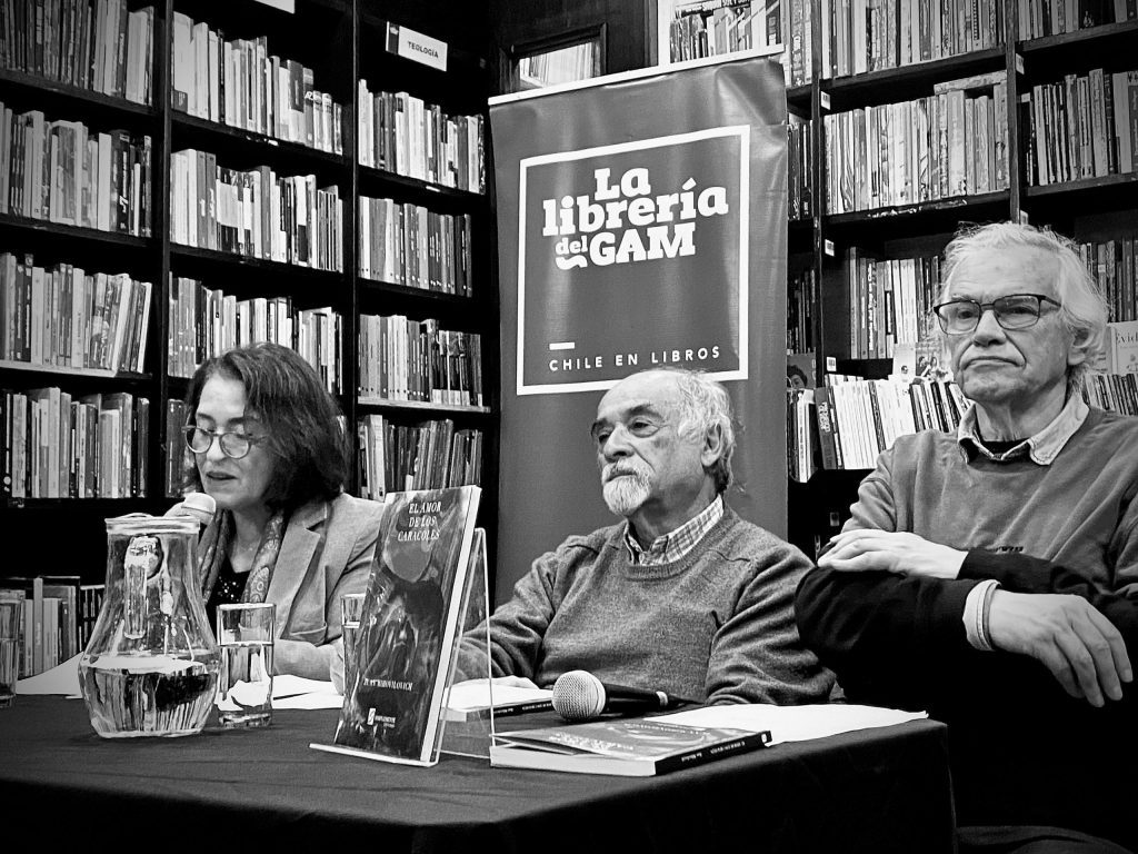 Tres personas en panel de literatura en biblioteca, Juan Mihovilovich al centro, con libros y cartel de evento al fondo