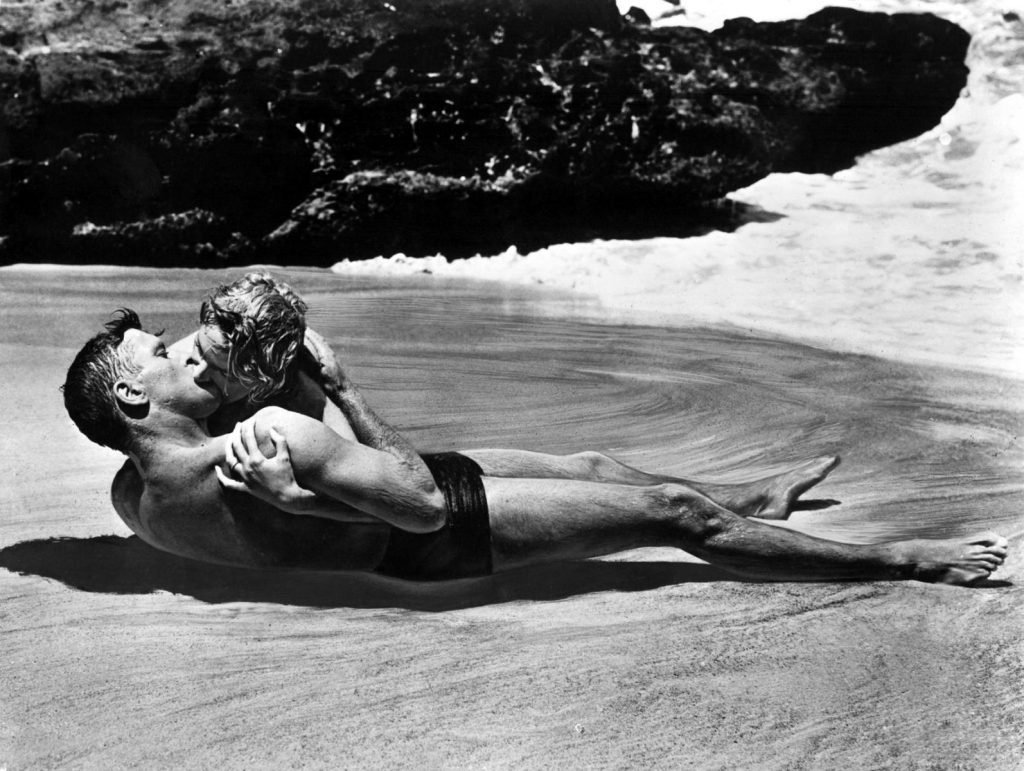 Pareja besándose en la playa junto a las rocas, escena icónica de romance en el cine clásico