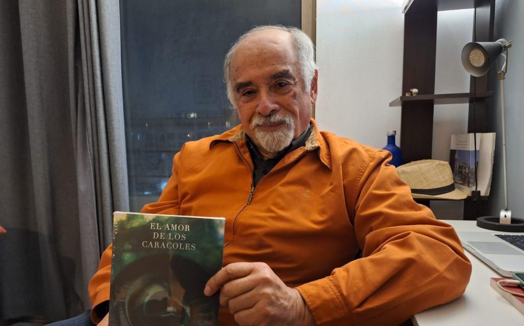 Escritor Juan Mihovilovich sostiene el libro El amor de los caracoles, vistiendo camisa naranja, en ambiente de interior