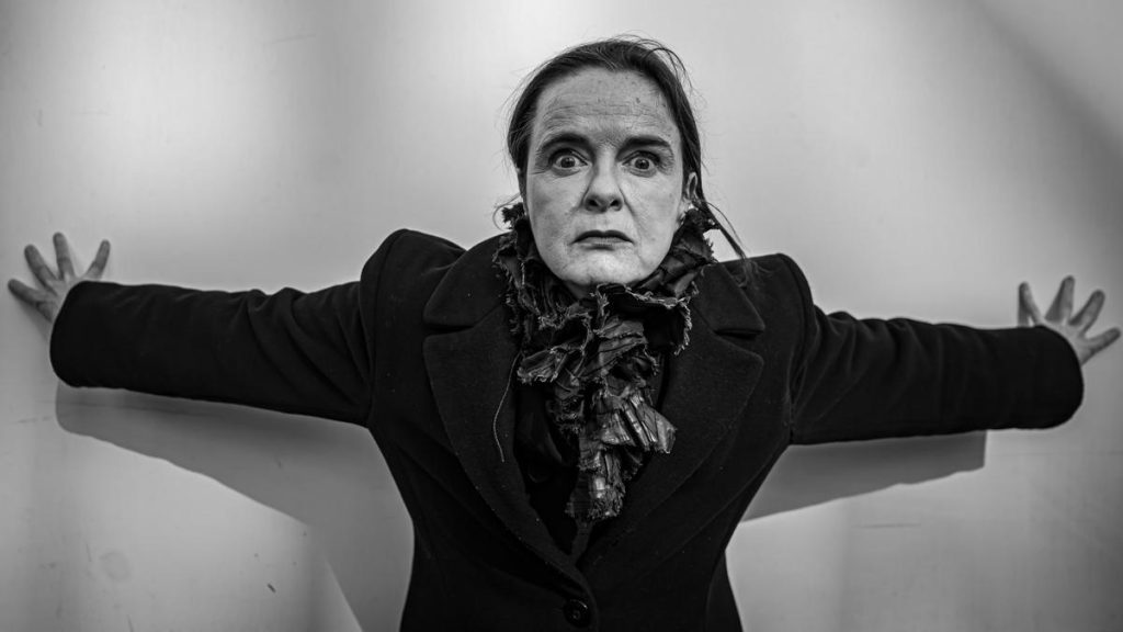 Amélie Nothomb con brazos extendidos en pose teatral, fotografía en blanco y negro, expresión seria y mirada frontal