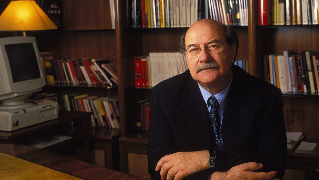 Antonio Skármeta en su oficina, sentado ante escritorio con biblioteca al fondo y lámpara dorada