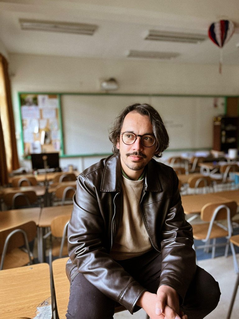 Cristian Rodríguez sentado en aula vacía con escritorios de madera, vistiendo chaqueta negra y gafas