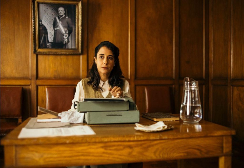 Mujer sentada tras escritorio de madera con libros apilados, cuadro al fondo, interior de oficina clásica