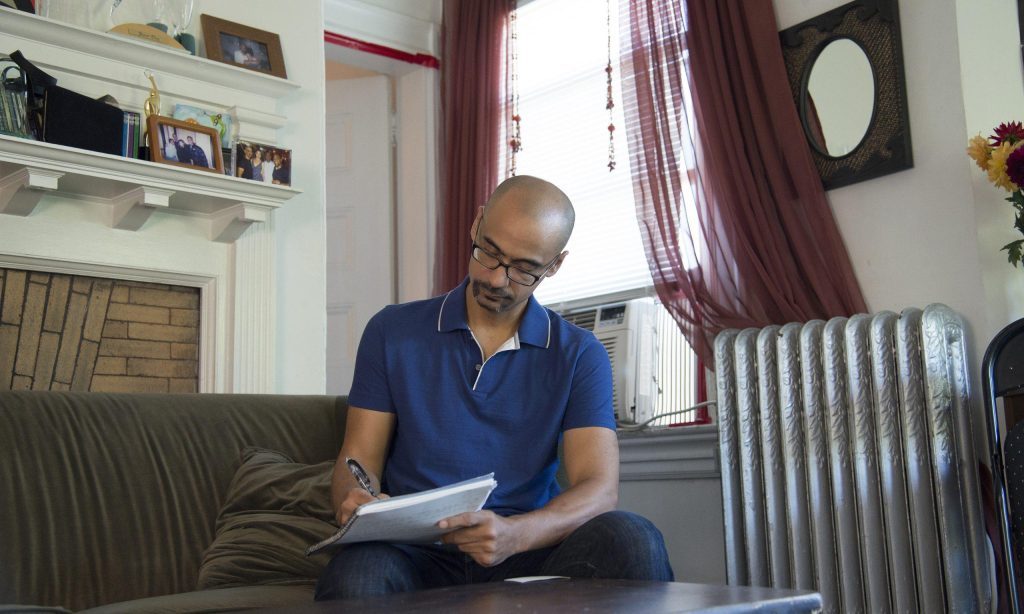 Junot Díaz, escritor dominicano, sostiene un libro mientras lee sentado en un sofá junto a una ventana con cortinas