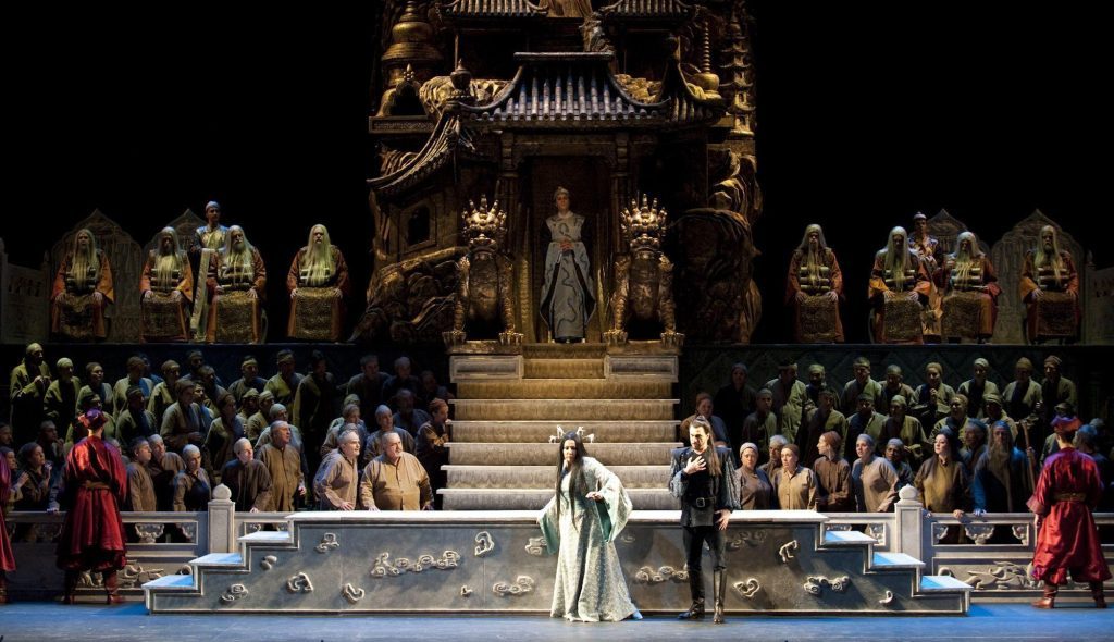 Escena de la ópera Turandot con protagonista en vestido blanco en el centro del escenario dorado, rodeada de coro