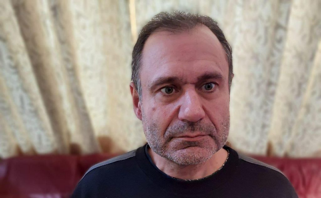 Aníbal Ricci Anduaga, hombre con cabello gris, expresión seria, sentado frente a cortinas beige