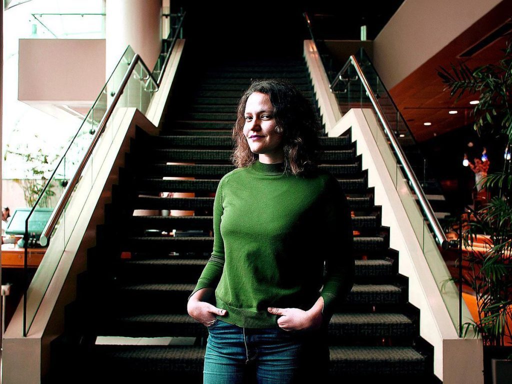 Mujer con cabello largo y oscuro, vistiendo suéter verde, de pie en escaleras de edificio interior