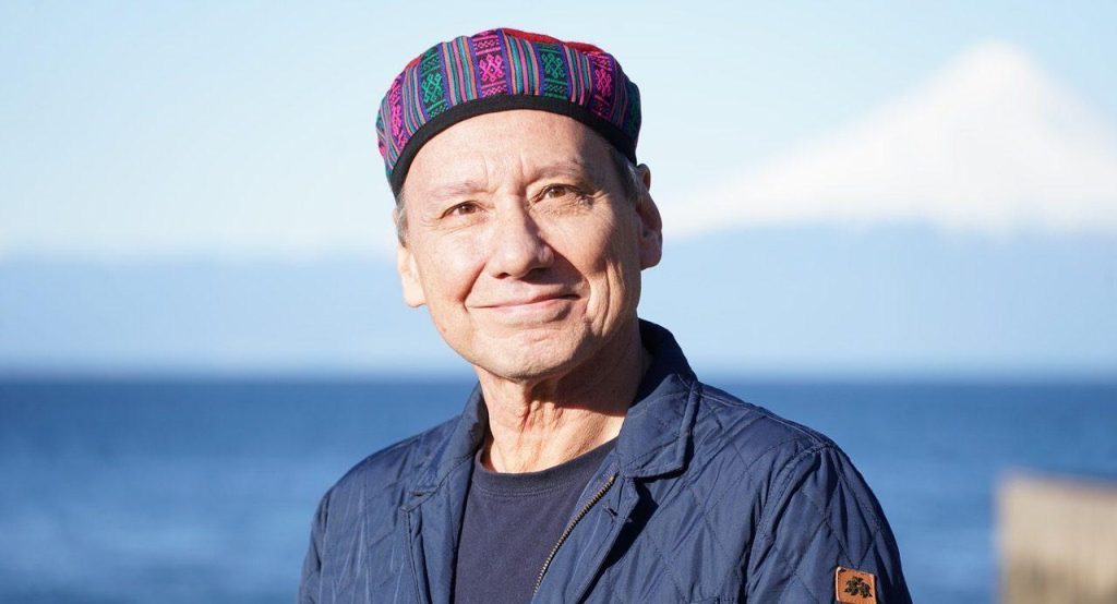 Claudio Suárez sonríe mirando a cámara, vistiendo gorra escocesa y chaqueta azul oscura, con el mar de fondo