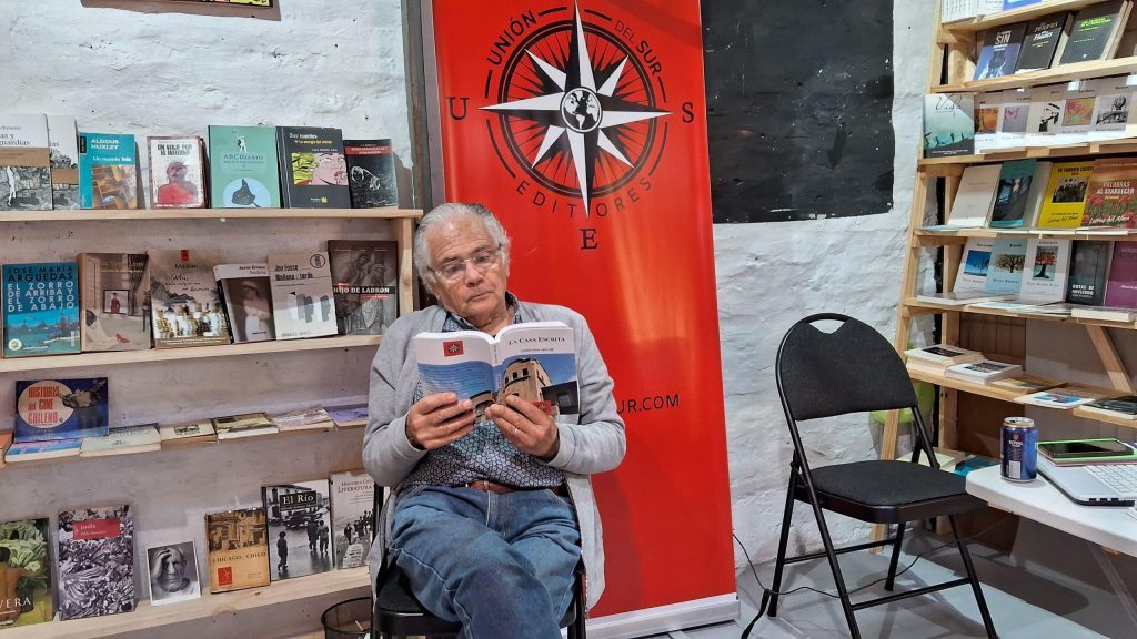 Edmundo Moure Rojas sostiene un libro en una sala con estanterías de literatura, frente a una pared roja con símbolo