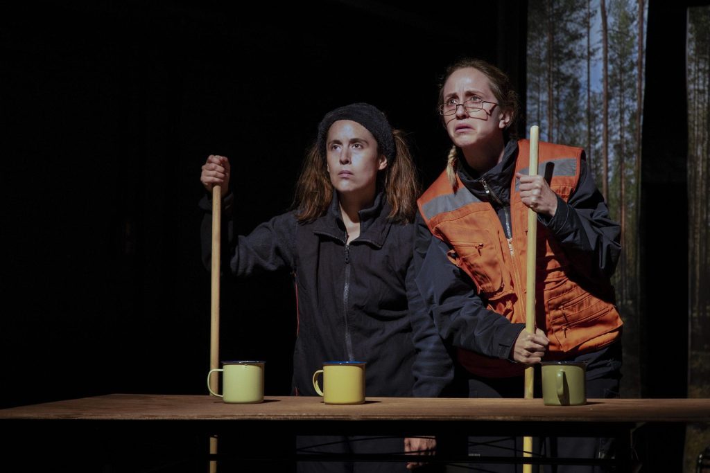 Tres actores en escena de teatro sobre tablado con hachas de madera y elementos de utilería, representando una obra