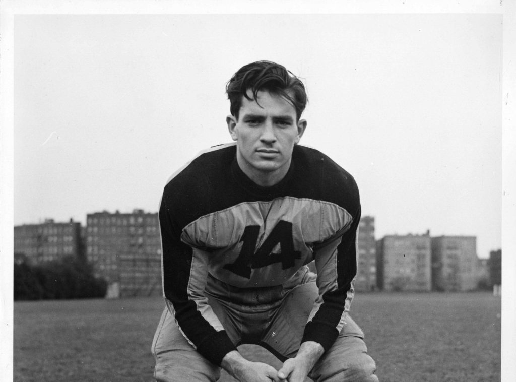 Jack Kerouac posando en actitud relajada sobre el césped, vistiendo jersey oscuro con letra, fondo urbano con edificios