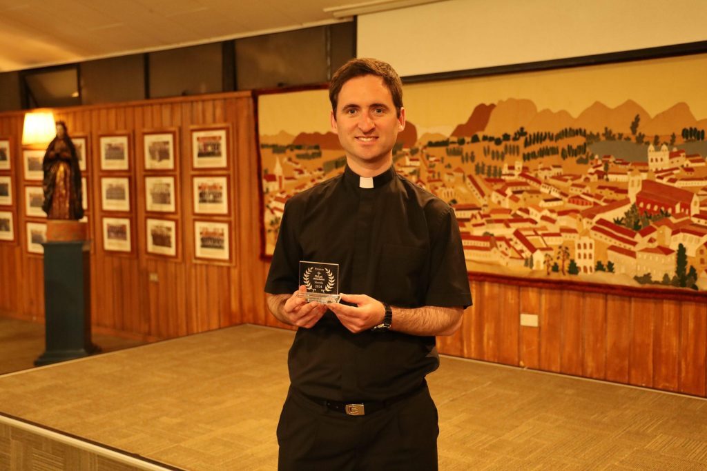 Juan Ignacio Izquierdo en galería con exposición de arte, sosteniendo premio literario, vistiendo sotana negra