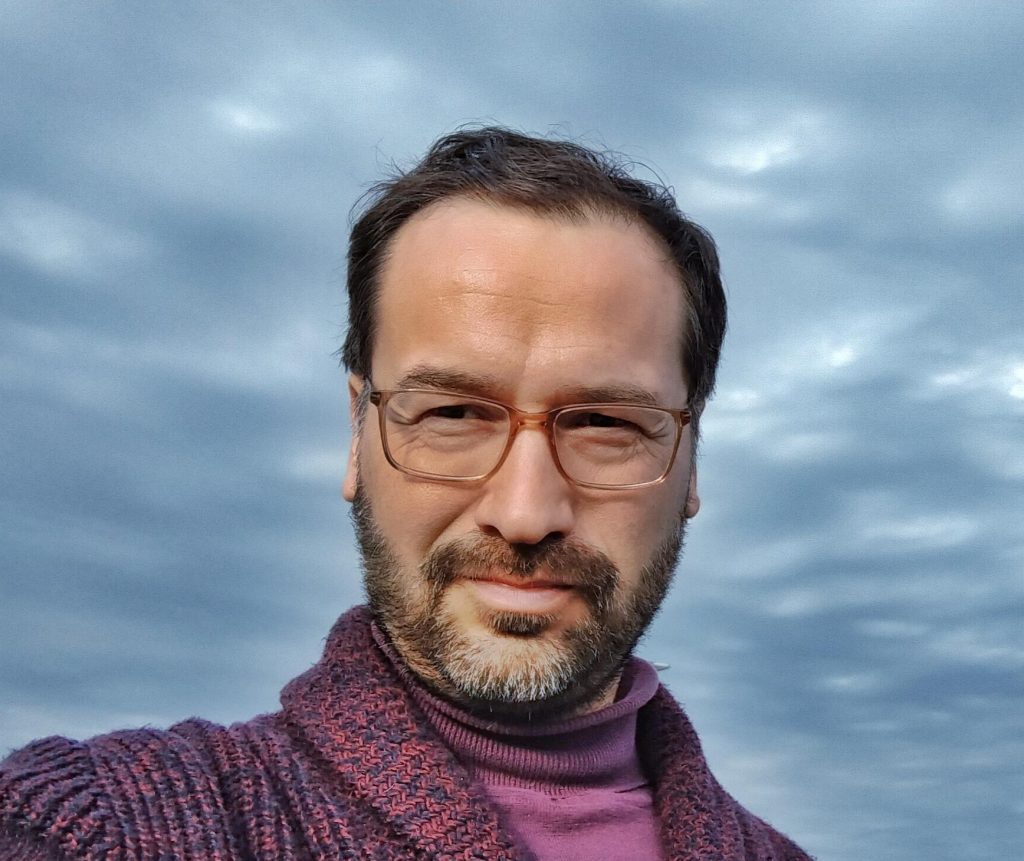 Luis Cruz-Villalobos, hombre con gafas y barba, sonriendo ante cielo nublado