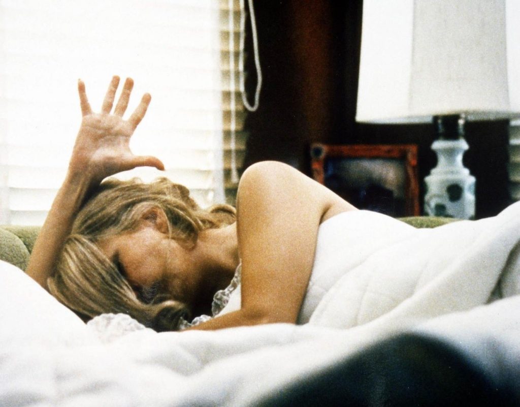Mujer en cama con expresión de angustia, mano en la frente, ambiente doméstico desordenado