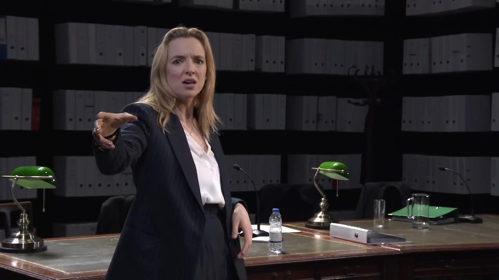 Mujer en traje oscuro gesticula expresivamente en oficina moderna con estanterías de fondo