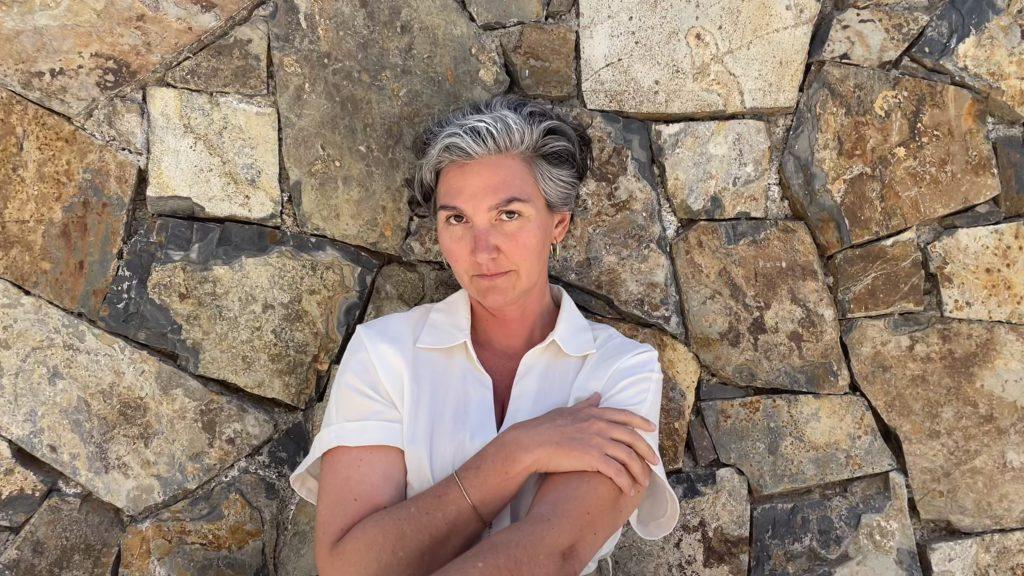 Mujer de cabello gris corto en blusa blanca, brazos cruzados, frente a pared de piedra