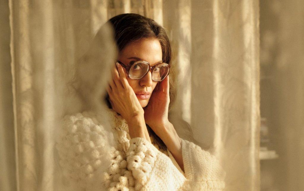 Mujer de cabello oscuro y lentes, con expresión pensativa, apoyando su rostro en las manos frente a cortinas doradas