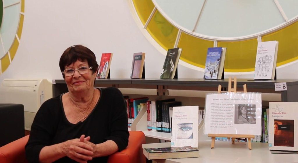 Nelly Lemus en su espacio de trabajo, rodeada de libros y materiales culturales, sonriendo hacia la cámara