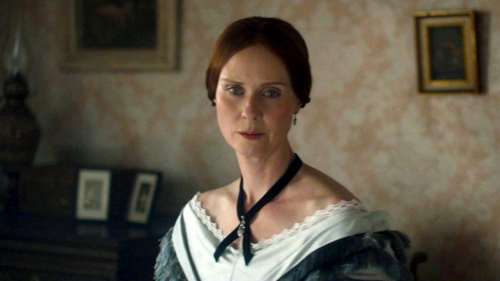 Mujer de época victoriana con vestido oscuro y collar blanco, retrato en interior con papel tapiz y marcos al fondo