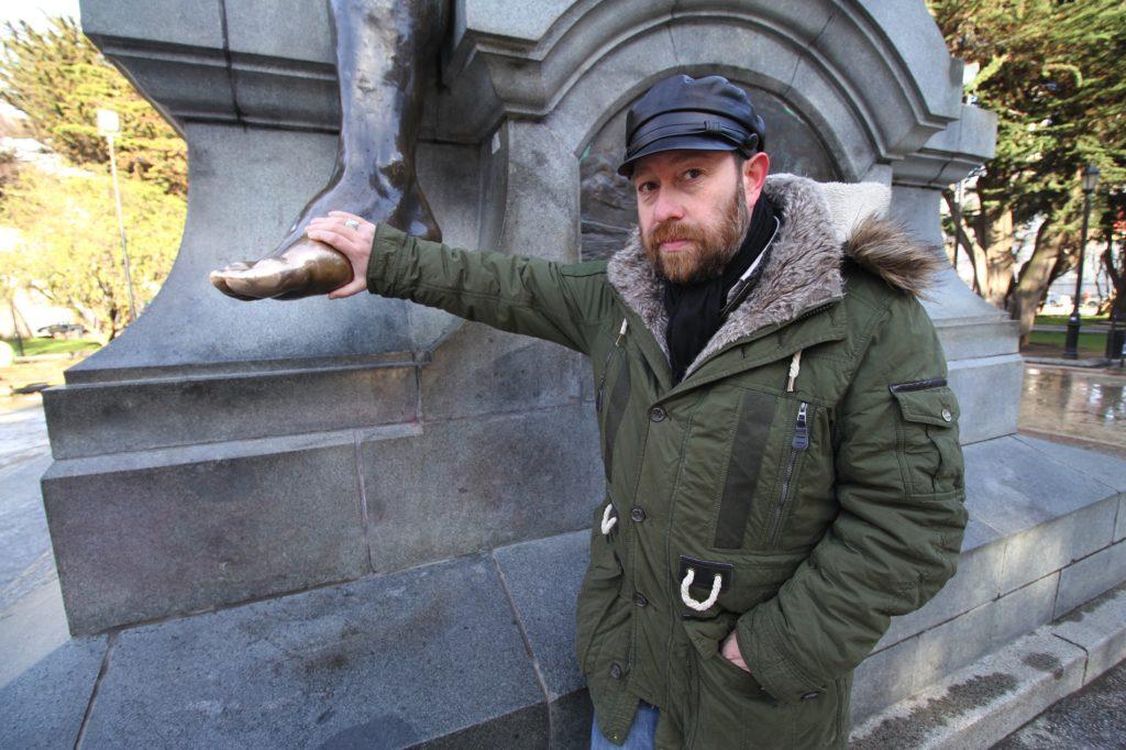 Óscar Barrientos junto a una estatua de piedra, extendiendo el brazo en gesto teatral, vistiendo abrigo verde oliva