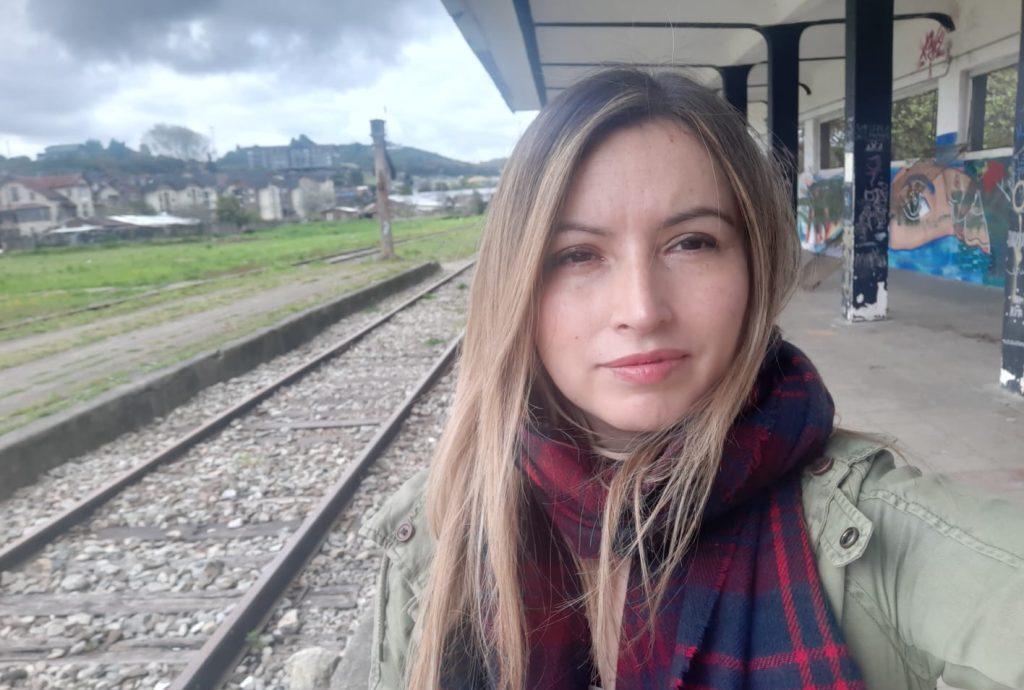 Mujer joven con cabello largo castaño y bufanda a cuadros, sonriendo en andén de estación de tren