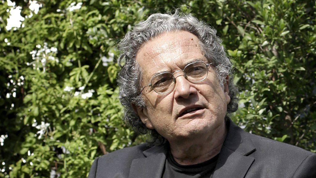 Escritor de mediana edad con gafas y cabello gris, vistiendo chaqueta negra, posando frente a follaje verde